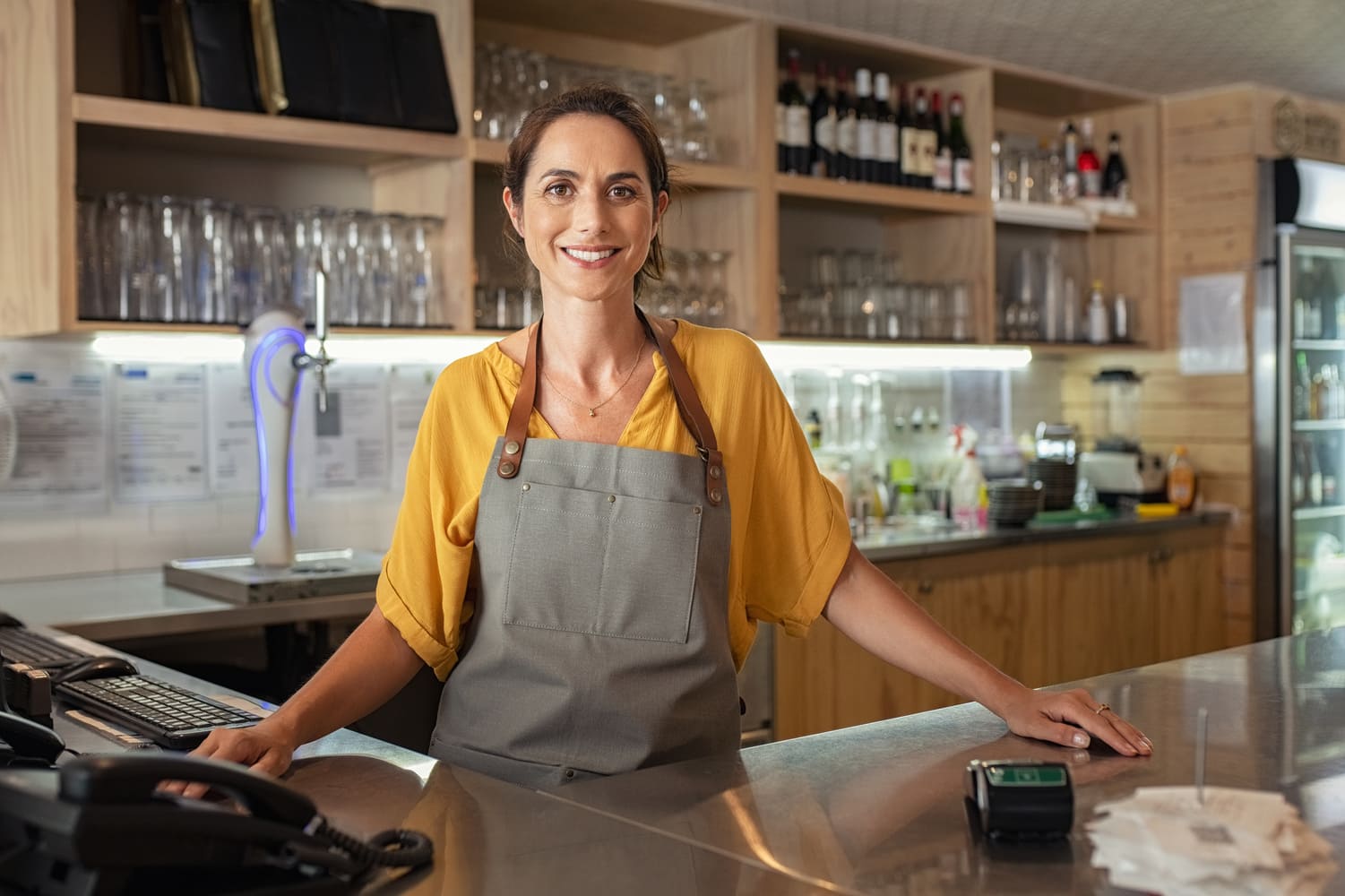 Mature woman barista standing behind the bar counter in coffee shop and looking at camera. Smiling small business owner smiling behind the counter. Portrait of beautiful waitress wearing apron.